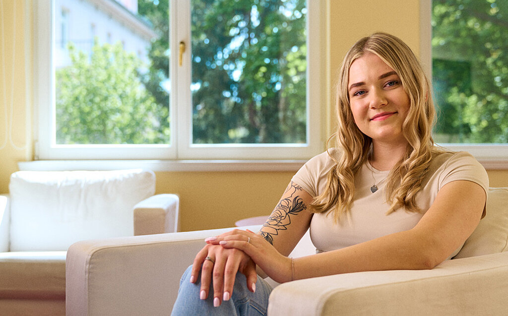 Porträt von Lilly, einer jungen blonden Frau, vor grauem Hintergrund.